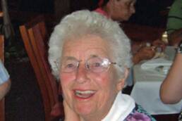 Smiling elderly woman wearing glasses with people seated at a dining table in the background