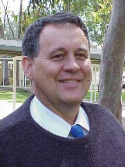 Headshot of an older man wearing a brown jumper, white shirt and tie with a tree and house in the background