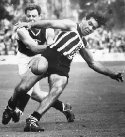 Man in a striped jersey kicking a football during an Australian Rules football game as another man runs behind him