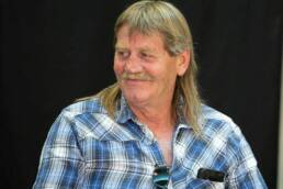 Headshot of a smiling man with shoulder length blonde hair and a mustache wearing a blue plaid shirt