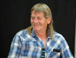 Headshot of a smiling man with shoulder length blonde hair and a mustache wearing a blue plaid shirt