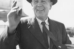 Man in a suit and top hat smiling and waving with an airplane in the background