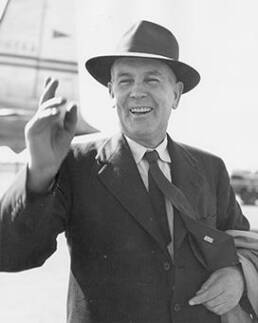 Man in a suit and top hat smiling and waving with an airplane in the background