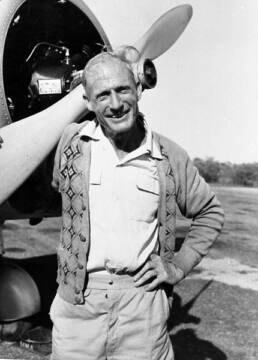 A smiling bald man standing in front of an airplane with one hand holding a propeller and the other on his hip