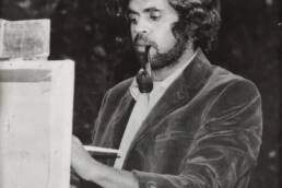 Curly-haired man with a beard smoking a pipe and painting at an easel