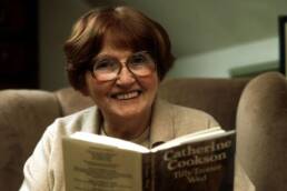 A smiling older woman with brown hair in a bob and glasses holding up an open book