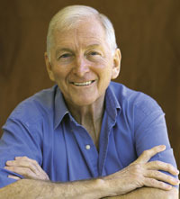 Headshot of an elderly man in a blue button-up shirt with arms crossed smiling at the camera