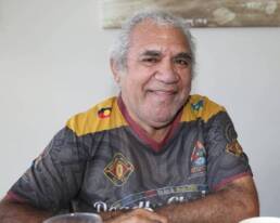 Smiling grey-haired man seated and wearing a grey and yellow shirt with a small Aboriginal flag logo