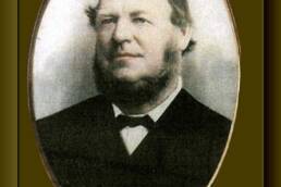 Headshot in a gold oval frame of a man with a full beard and comb-over hairstyle wearing a black suit