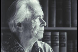 Profile headshot of a white-haired man in a wide collared shirt with books in the background