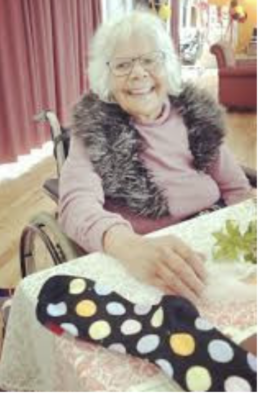 Smiling woman with white hair and glasses sitting in a wheelchair at a table.