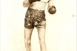 Man with short hair wearing black shorts, boxing gloves and no shirt standing in with fists up in a boxing pose