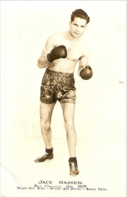 Man with short hair wearing black shorts, boxing gloves and no shirt standing in with fists up in a boxing pose
