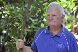White haired man wearing a blue polo shirt with a small Australian flag near the collar with his hand wrapped around a small tree