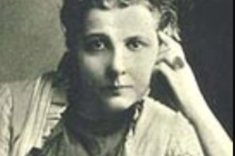 Headshot of a woman with very short hair wearing a white jacket and loose blouse with her hand resting on the side of her face