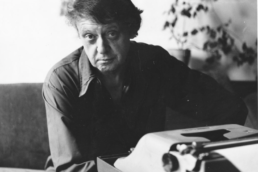 A bushy-haired man in a button up shirt seated in front of a typewriter with a houseplant in the background