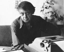 A bushy-haired man in a button up shirt seated in front of a typewriter with a houseplant in the background