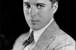 Black and white headshot of a young man with wavy hair wearing a tweed suit and seated at an angle