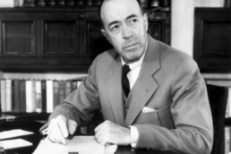 Black and white headshot of a balding man in a suit seated at a desk with an ornate bookshelf in the background