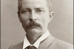 Headshot of a mustached-man with a sombre expression wearing a suit