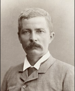 Headshot of a mustached-man with a sombre expression wearing a suit