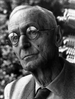 Headshot of an older man with round eyeglasses wearing a tweed suit with a neutral expression