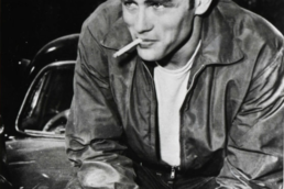 Black and white image of a young man with a mischievous smile sitting on a car with a cigarette in his mouth