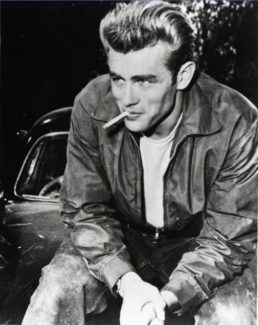 Black and white image of a young man with a mischievous smile sitting on a car with a cigarette in his mouth