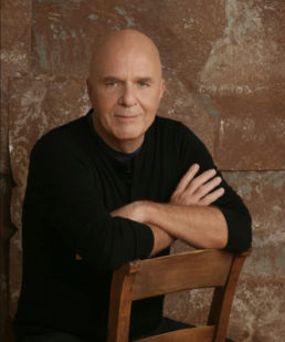 A bald man wearing a black shirt sitting backwards on a wooden chair against a brick wall