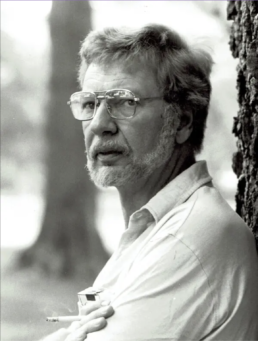 Black and white headshot of a grey haired man with beard and glasses smoking a cigarette next to a tree.