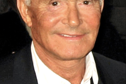 Headshot of a smiling grey haired man with dark eyes wearing a white collared shirt and jacket