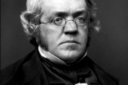 Black and white headshot of a grey-haired man with mutton chops wearing glasses and a suit