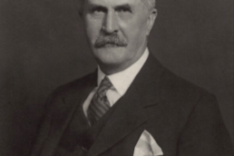 Black and white headshot of a balding older man with a mustache wearing a suit, waistcoat and handerchieve