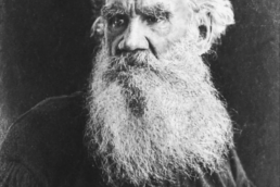 Black and white headshot of a balding elderly man with a long beard and mustache wearing a black button-up shirt