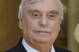 Headshot of an elderly man with a mustache wearing a suit and maroon tie