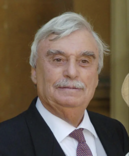 Headshot of an elderly man with a mustache wearing a suit and maroon tie