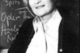 Headshot of an older woman wearing a button-up shirt and cardigan in front of a chalkboard