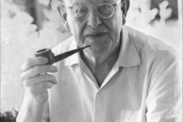 Black and white headshot of a bald older man with glasses in a white shirt holding a pipe