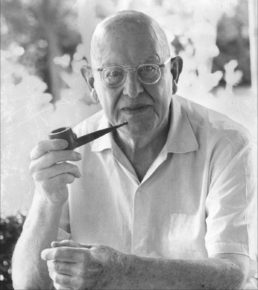 Black and white headshot of a bald older man with glasses in a white shirt holding a pipe