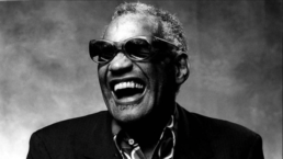 Black and white headshot of a grey-haired man with an open-mouth smile wearing dark sunglasses, a button-up shirt and jacket