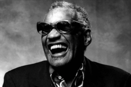 Black and white headshot of a grey-haired man with an open-mouth smile wearing dark sunglasses, a button-up shirt and jacket