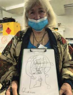 A grey haired woman wearing a face mask holding up a black and white drawing