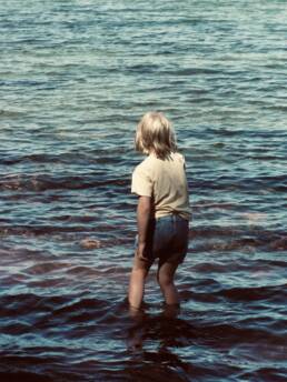 A young woman with blonde hair with her back to the camera knee deep in the ocean.