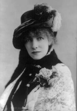 Black and white headshot of a woman in a lacey dress and wide-brimmed hat with a corsage.