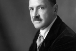 Black and white headshot of a middle-aged man with crossed arms, a mustache and dark slicked-back hair