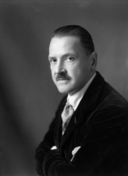 Black and white headshot of a middle-aged man with crossed arms, a mustache and dark slicked-back hair
