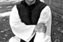 Black and white image of a bald man in a cassock resting his hand on one arm in front of a building