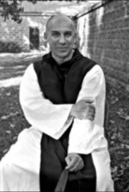 Black and white image of a bald man in a cassock resting his hand on one arm in front of a building