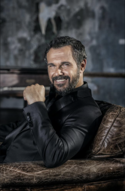 Smiling middle-aged man with a beard sitting on a leather sofa wearing a black collared shirt
