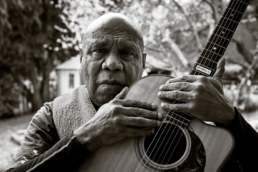 Black and white headshot of an older man holding a guitar with a house and trees in the background